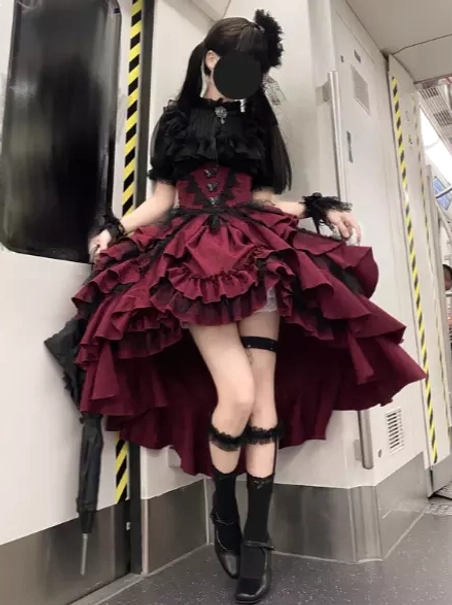 Gothic lolita dress set front three-quarter view in subway carriage featuring ruffled high-low skirt, black lace wrist cuffs, black hair flower accessory, and leg garter styling