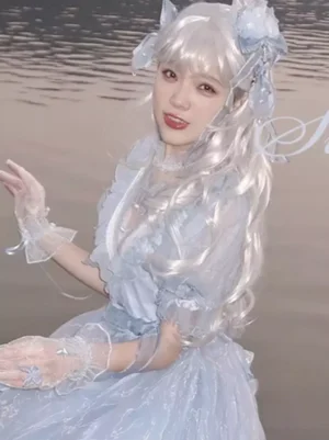 Silver long tousled curly Gothic Lolita wig with blunt bangs worn by model at waterside, featuring fluffy silvery-white waves, sheer lace gloves, and flower hair ornaments