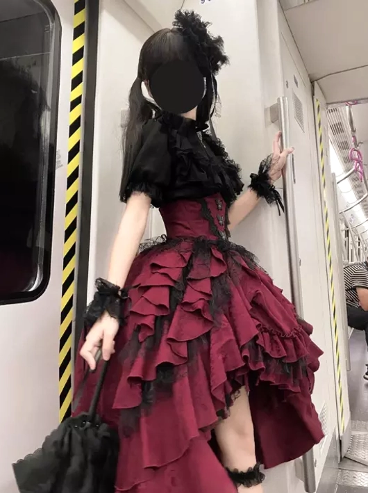 Gothic lolita outfit set back three-quarter view in subway carriage showing wine-red ruffled skirt with black blouse, black handbag, lace-trimmed cuffs, and complete styling details