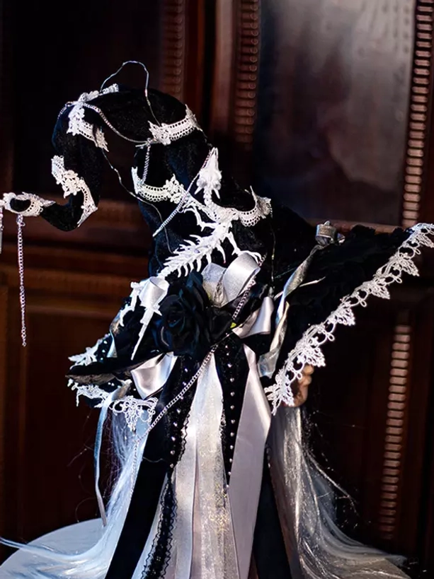 Left side three-quarter view of black and white gothic Lolita witch hat with silver long hair mannequin, featuring intricate lace trimmings, layered ribbon bows, and dangling metal chains on dark vintage wooden background