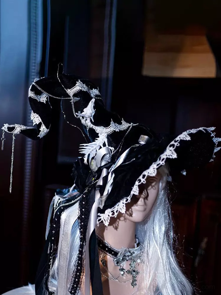 Low left side view of gothic witch hat headpiece worn on silver long-haired mannequin with coordinating crystal choker, black lace trims, and dangling decorative chains against dark wood indoor background