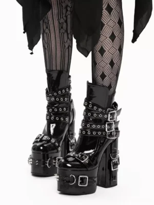Gothic platform ankle boots with metal grommets and patent leather, front oblique angle view showing glossy black finish with decorative buckle straps and thick block heel