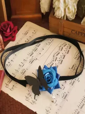Flat lay photography of Gothic rose eye patch mask with vintage sheet music, wooden rose box, featuring blue rose embellishment and black butterfly with dangling cross pendant on black velvet band