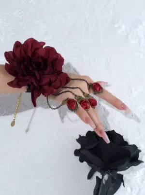 Gothic rose hand chain accessory set featuring large burgundy flower corsage with layered metal chain links, elevated top-down angle on white lace with dark fabric rose prop