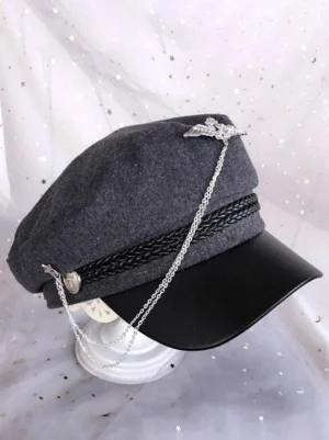Gray wool lolita military newsboy cap with cascading chain decoration and metal wing brooch, side three-quarter angle displayed on stand with starry tulle backdrop