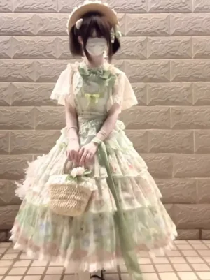 Green pastel floral gothic lolita JSK dress front full-body view with flutter short sleeves, prominent front bow, tiered lace skirt, worn with straw hat and woven flower basket on tiled background