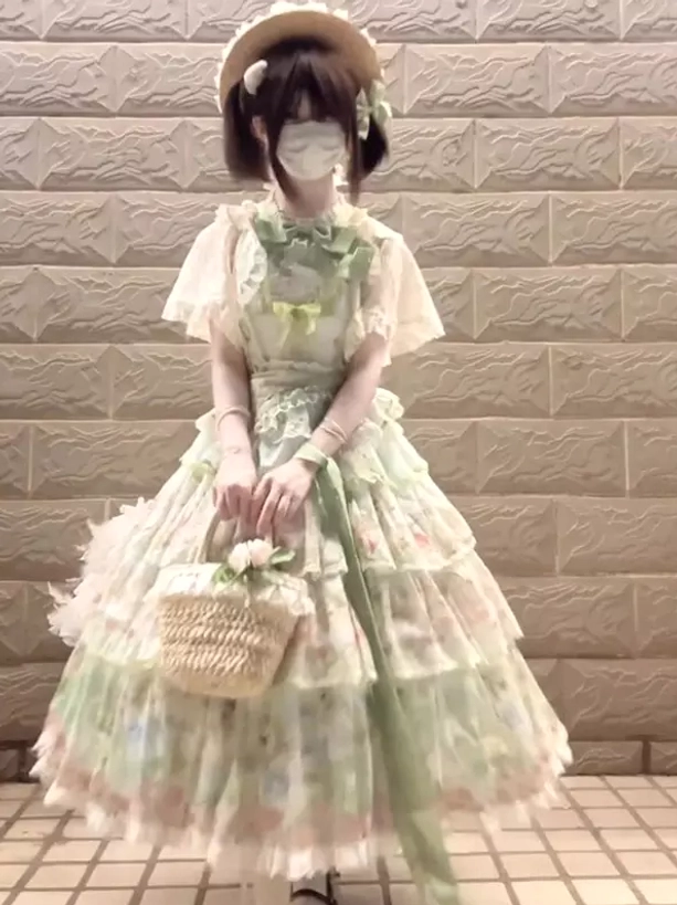 Green pastel floral gothic lolita JSK dress front full-body view with flutter short sleeves, prominent front bow, tiered lace skirt, worn with straw hat and woven flower basket on tiled background