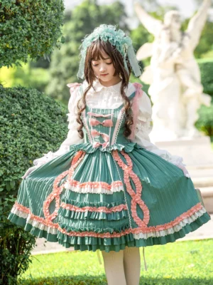 Green Rococo sweet lolita jumper dress front view with pink bows and lace, worn by model in garden setting with angel statues, featuring teal hair bonnet and white stockings