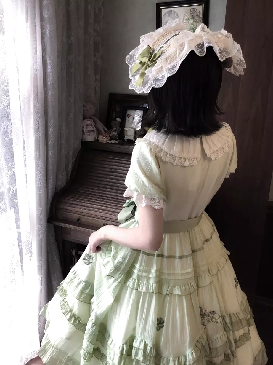 Back full-body view of green sweet lolita dress displaying tiered ruffled layers from behind, lace bonnet, ruffled collar detail, decorative waist sash, indoor room background