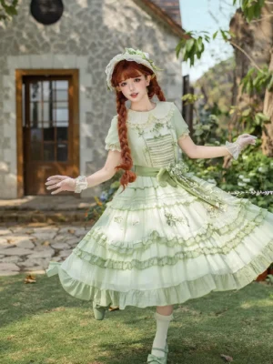 Full-body front view of green sweet lolita dress worn by model with red braids, outdoor garden setting, showing matching bonnet headwear, ruffled layered skirt, short puff sleeves, and Victorian-inspired silhouette