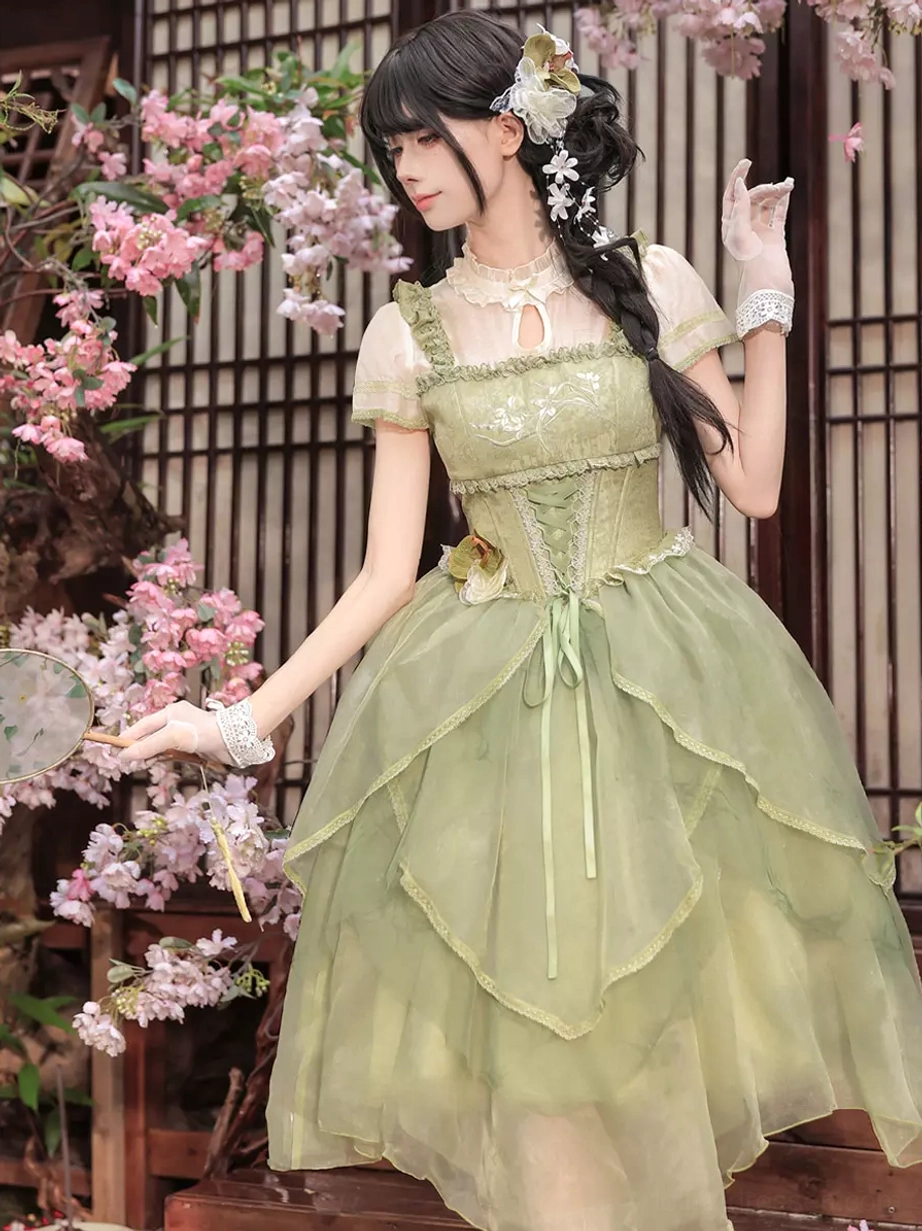 Front side view of green sweet lolita dress with sequin embellished bodice, wrist gloves, and asymmetrical tulle skirt with cherry blossom background aesthetic