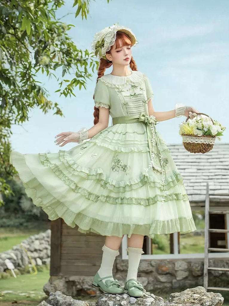 Full-body side view of green sweet lolita dress showcasing tiered ruffled skirt, model with red braids holding flower basket, wearing lace gloves and matching bonnet in countryside setting