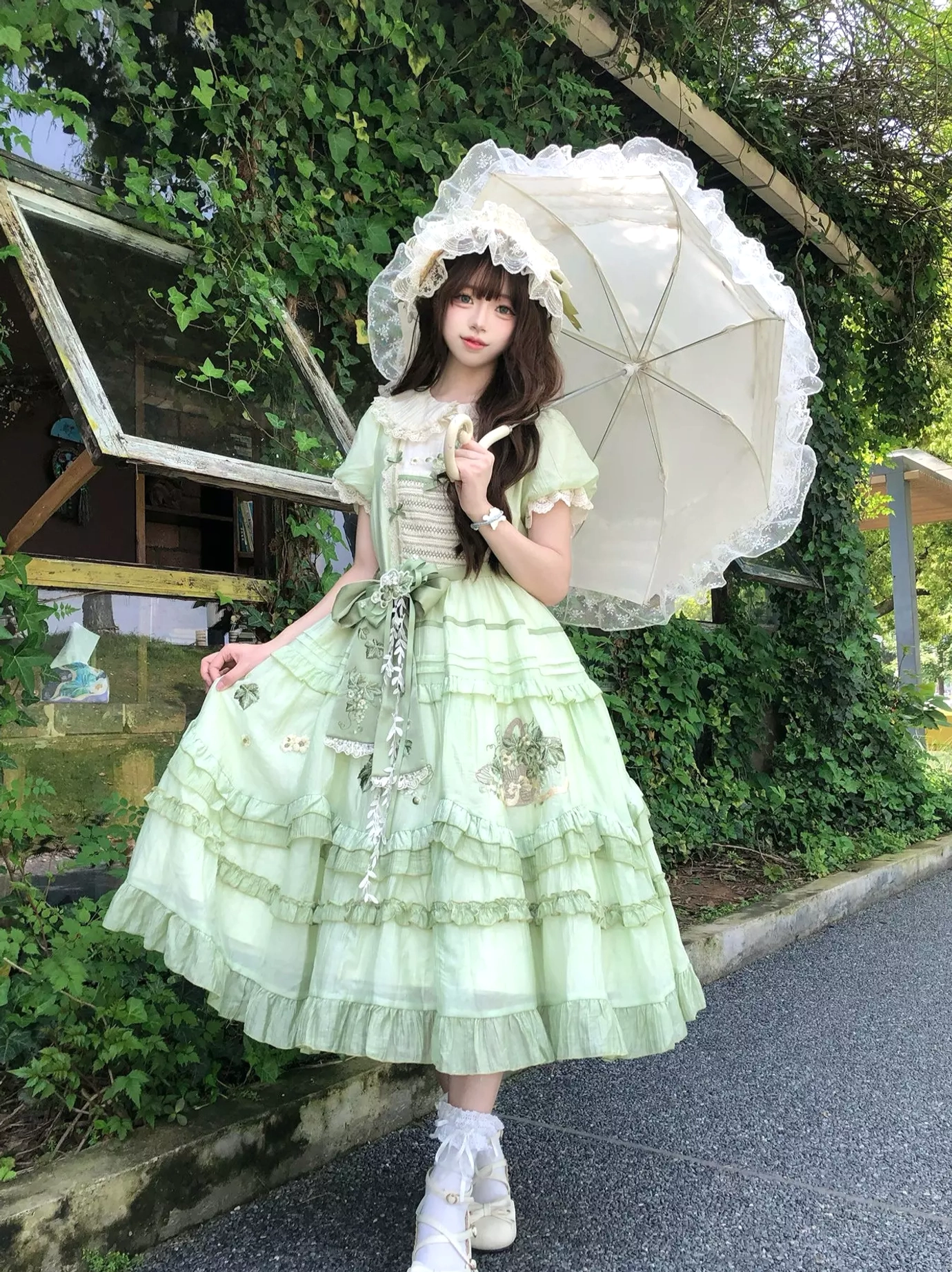 Three-quarter full-body view of green sweet lolita dress on model with dark hair, holding decorative lace parasol, wearing matching bonnet and embroidered skirt, greenery road background
