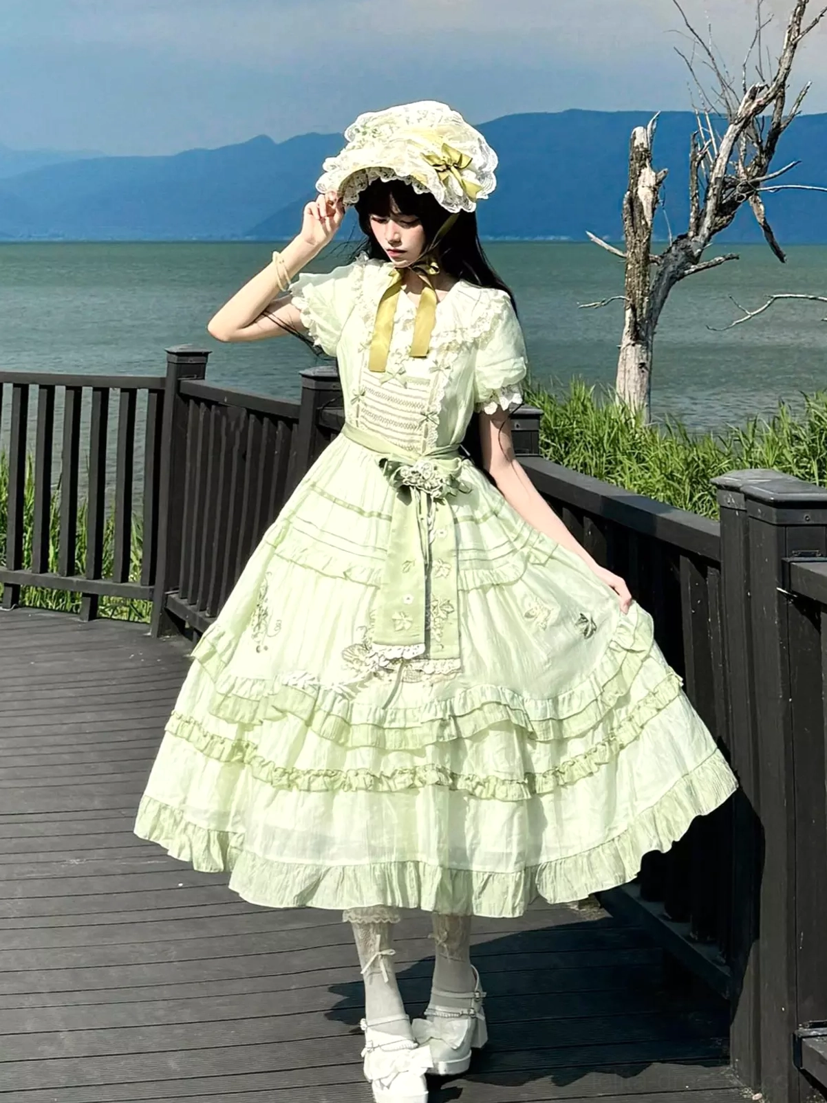 Full-body front view of green sweet lolita dress featuring matching bonnet, yellow neck bow accent, tiered embroidered skirt layers, model at lakeside wooden deck location