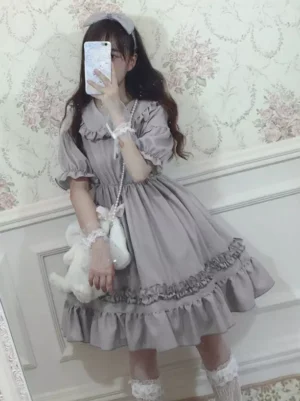Front close-up view of grey chiffon Sweet Lolita one-piece dress worn by model showing detailed ruffled layered skirt hem, Peter Pan collar embellishment, and lace wrist decorations on vintage wall background