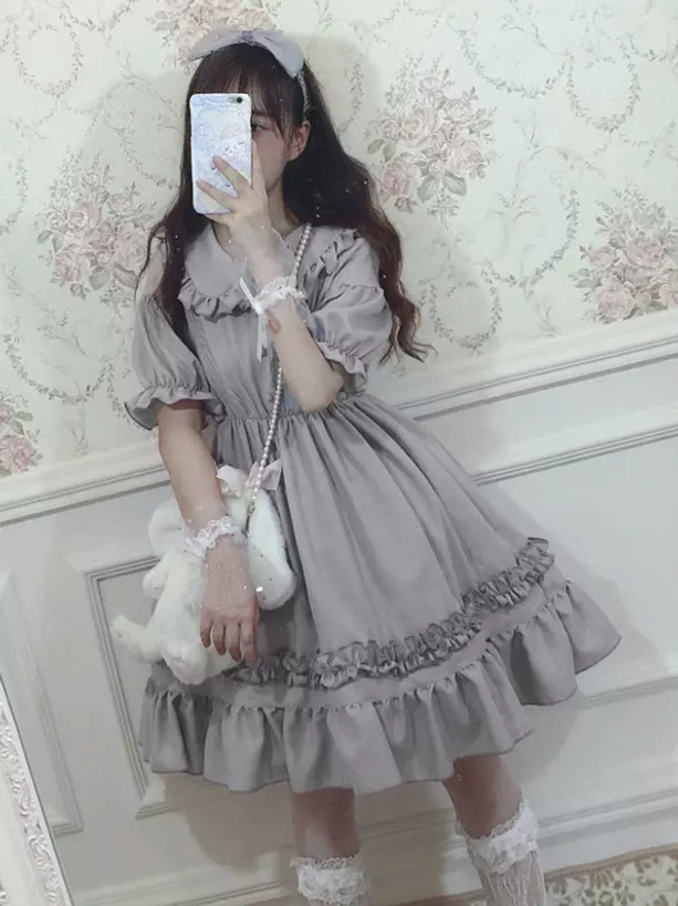 Front close-up view of grey chiffon Sweet Lolita one-piece dress worn by model showing detailed ruffled layered skirt hem, Peter Pan collar embellishment, and lace wrist decorations on vintage wall background