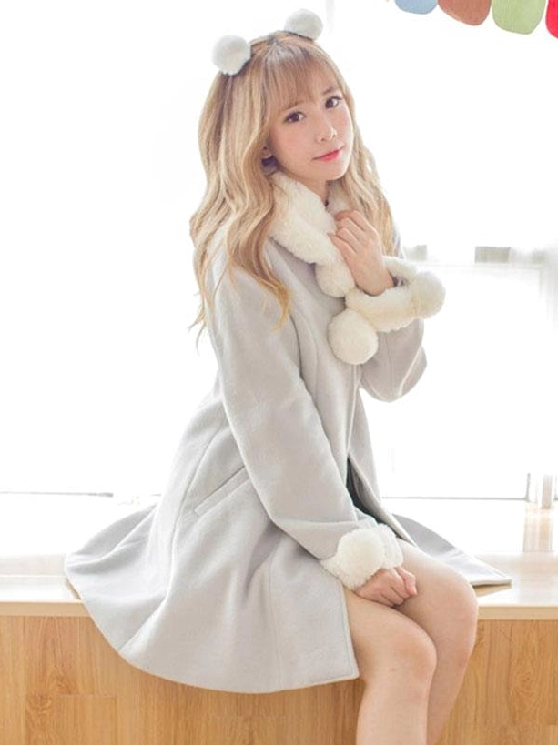 Side three-quarter view of girl sitting on wooden surface wearing grey wool lolita overcoat with white fuzzy collar, pom pom embellishments, and fuzzy cuffs, displaying the coat's volume, drape, and dimensional design details