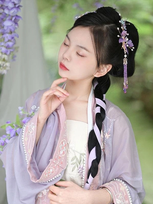Model wearing light purple hanfu with ancient braided hairstyle styled with side-mounted purple flower lolita hair ornament against outdoor wisteria flower background demonstrating authentic historical styling