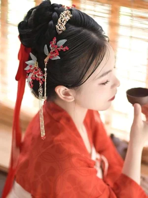 Woman wearing red Hanfu dress styled with gold chain tassel hair accessories and red berry ornaments, holding traditional tea bowl, side profile view showcasing delicate hair comb and frosted blue leaf details
