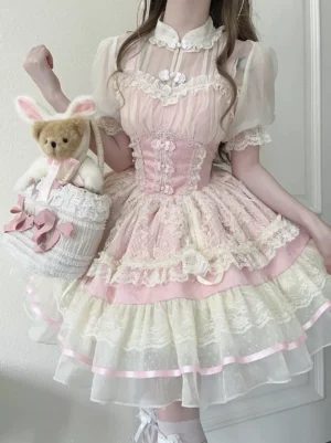 Front three-quarter view of blush pink Hime Lolita OP dress with tiered lace ruffles, pleated puff sleeves, and structured corset waist, worn by model with decorative accessories in home setting