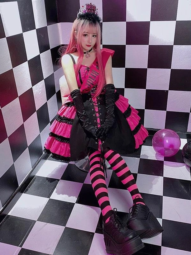hot pink gothic sweet lolita jsk dress side sitting view with pink-haired model wearing black gloves striped stockings tiered ruffled skirt