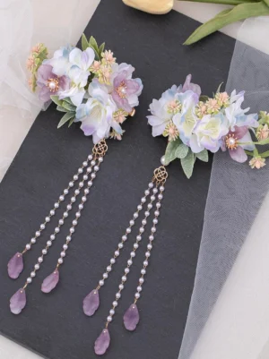 Lavender floral tassel hair clips pair flat lay top-down view featuring purple-blue artificial blooms with pearl centers, triple-strand beaded tassels with teardrop purple pendants, square hollow metal connectors, and gold hair clip bases on black slate background