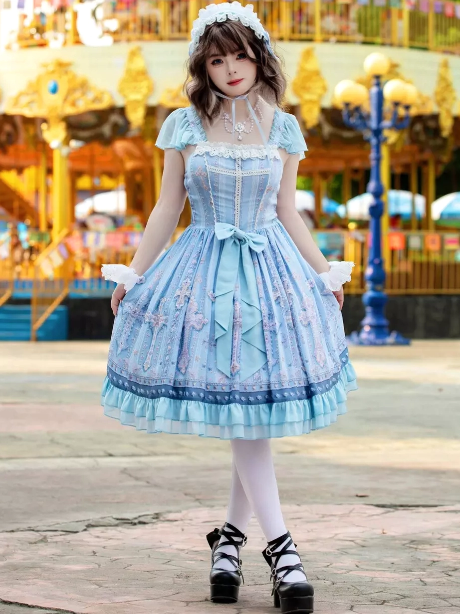 Light blue cross print sweet lolita JSK dress full front view with ruffled cap sleeves, square lace neckline, fitted waist bow, and all-over cross pattern print on model at amusement park carousel background