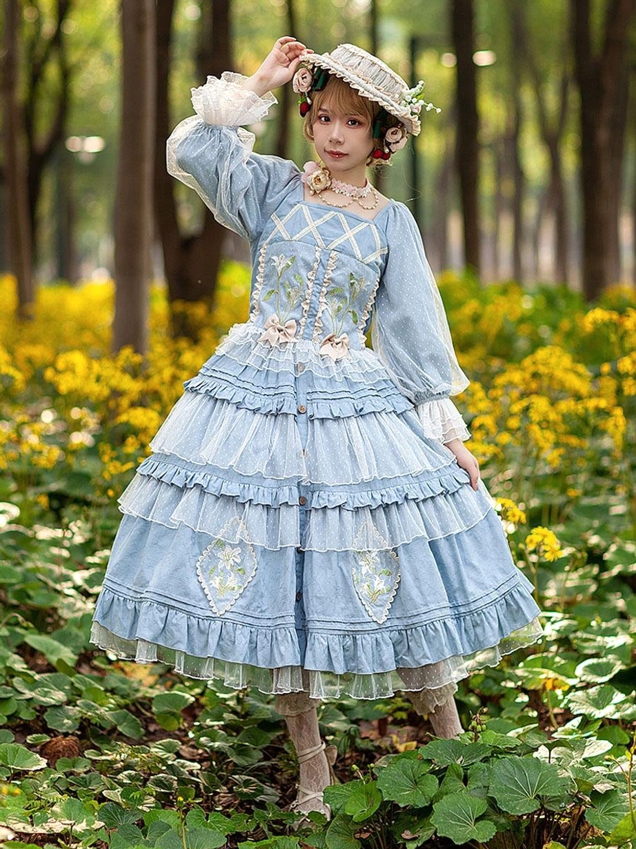 Light blue Classic Lolita dress full-body front view on model wearing straw hat in woodland flower field showing square neckline puff sleeves and floral embroidery