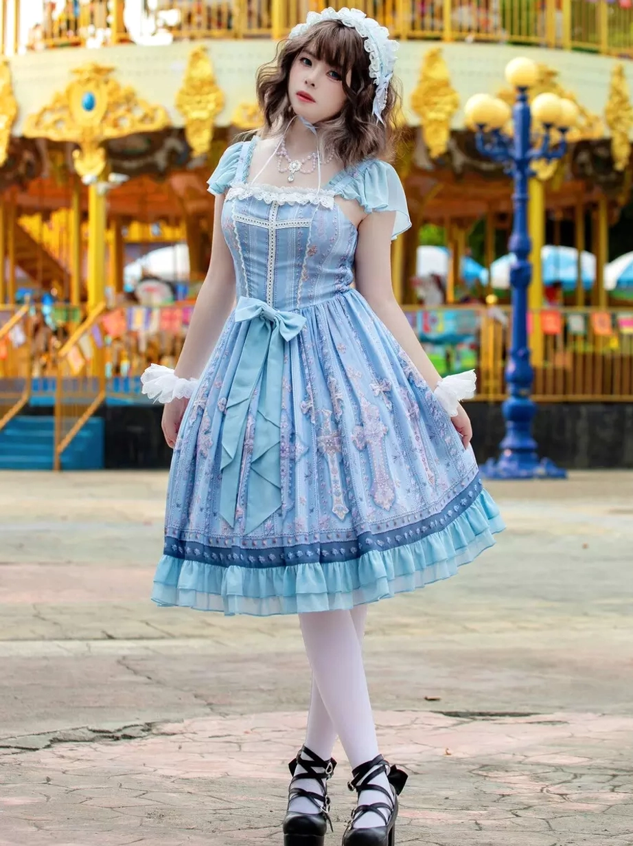 Light blue lolita dress angled full front view featuring lace-trimmed cap sleeves, all-over cross print pattern, fitted waist bow, ruffled A-line skirt against amusement park background