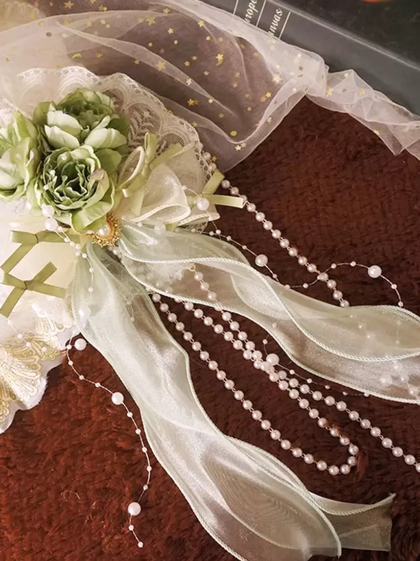 Side profile view of light green lolita hair accessory displaying delicate construction with sheer ribbons, green artificial flowers, pearl chain details and intricate lace trim on starry tulle background with vintage props