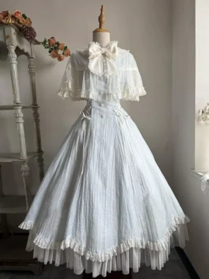 Light sky blue sweet lolita jumper skirt dress full-length front view with cream big bow, lace cape shawl, double-layer ruffled hem, and vertical pleating displayed indoors with floral decorations