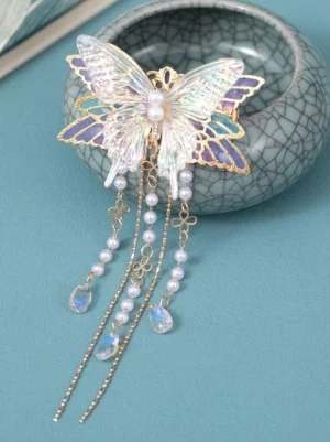 Lilac iridescent butterfly hair tassel accessory with pearl and chain details displayed on cracked ceramic vessel, showcasing the shimmering resin butterfly wings and gold-edged purple-blue accents from front top-down perspective for lolita fashion styling