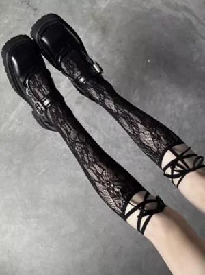 Black lace knee-high socks with delicate floral crochet pattern and cross-tie ankle fasteners styled with black platform Mary Jane shoes on concrete ground, overhead shot showing intricate lace texture and Lolita fashion aesthetic