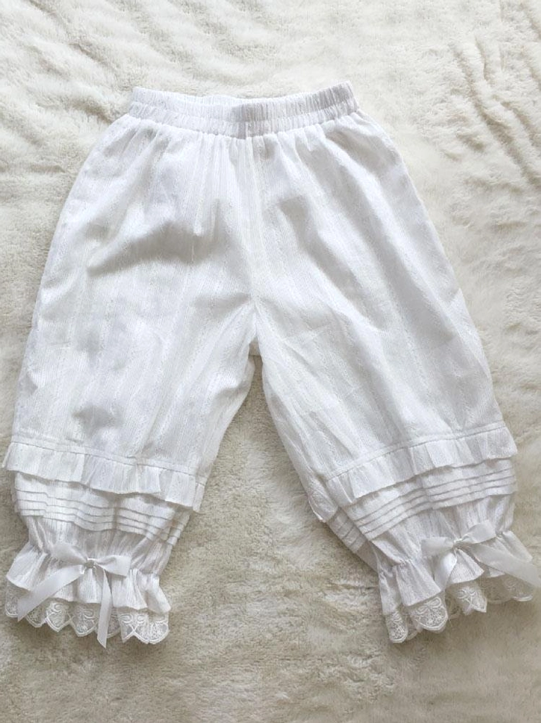 White lolita bloomers detail shot showing satin bow cuff embellishments, ruffled lace hem construction, and vertical striped jacquard texture, overhead flat lay photography