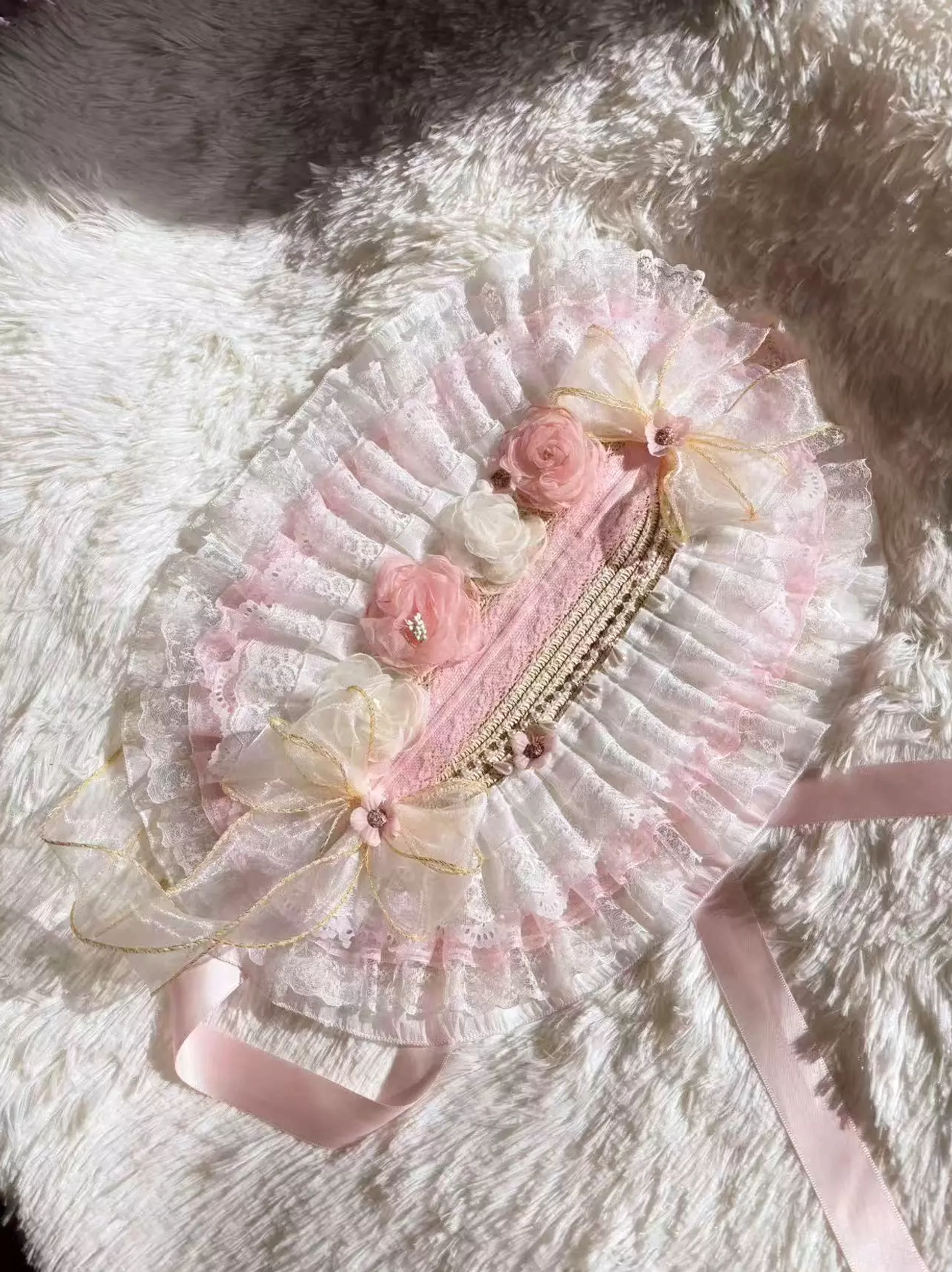 Flat lay top-down view of sweet lolita bonnet headdress displaying ruffled lace construction, artificial pink roses, sheer gold floral ornaments, and pink satin chin straps on white fur background