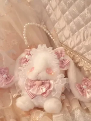 Mini Lolita style rabbit plush accessory bag with soft pink gingham bows, pearl handle detail, adorable blush cheeks, flat lay presentation on quilted lace fabric background