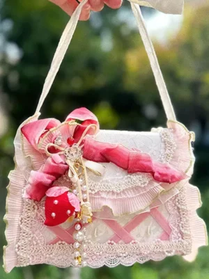 Sweet lolita handbag flat front view with pink ribbon bow, lace ruffle details, strawberry beaded tassel, and pink cross webbing design outdoor foliage background