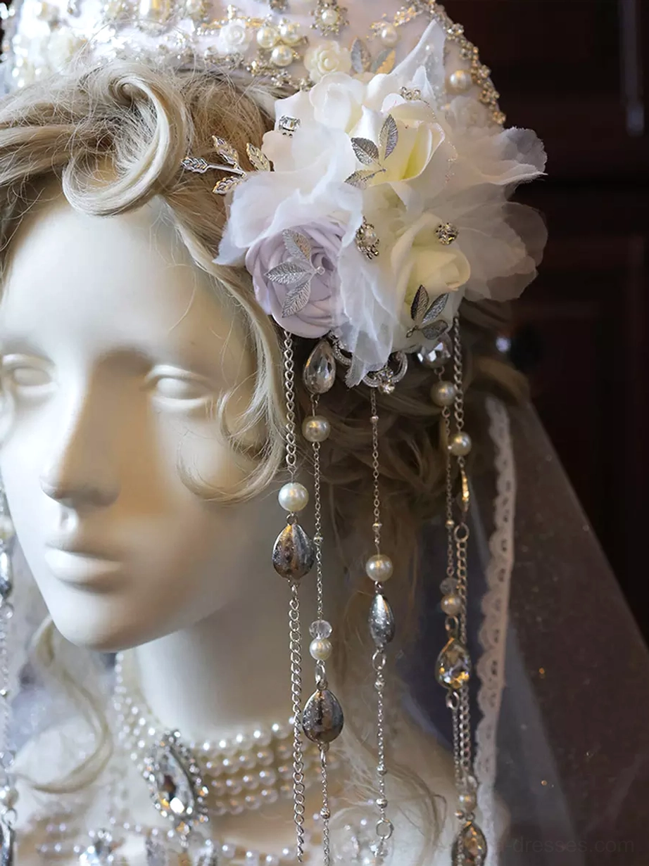 Detailed close-up view of lolita bridal crown showing large white purple fabric flowers with diamond embellishments, multiple pearl and crystal drop chains, ornate metal framework, and chain jewelry details