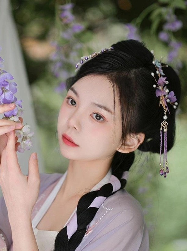 Front portrait view of hanfu-dressed model with elaborate braided hairstyle wearing multiple purple flower lolita hair ornaments on sides and top of head holding wisteria flowers showing complete coordinate styling