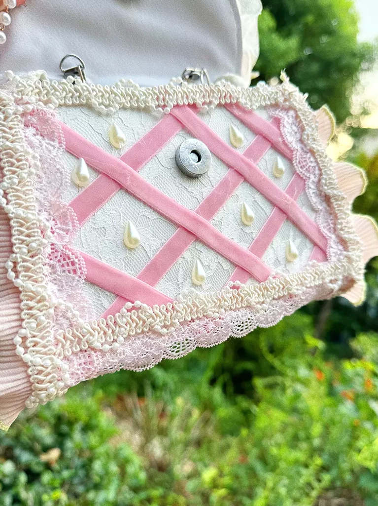 Lolita handbag bottom panel overhead view showing pink cross webbing, lace trim details, metal snap buckle, and water drop decorative patterns in garden background