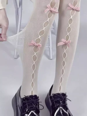 Lolita hollow mesh pantyhose with vertical cutout details and bow embellishments, close-up view of lower legs and feet wearing black Mary Jane shoes, showing beige base with pink bow accents and stretchable mesh construction