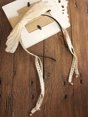 Delicate lace katyusha headband flat lay overhead view on dark brown wooden surface, displaying black thin hair band base, off-white intricate lace details, ruffled bow construction, and tiny pearl charms positioned on ribbon ends for lolita fashion accessory