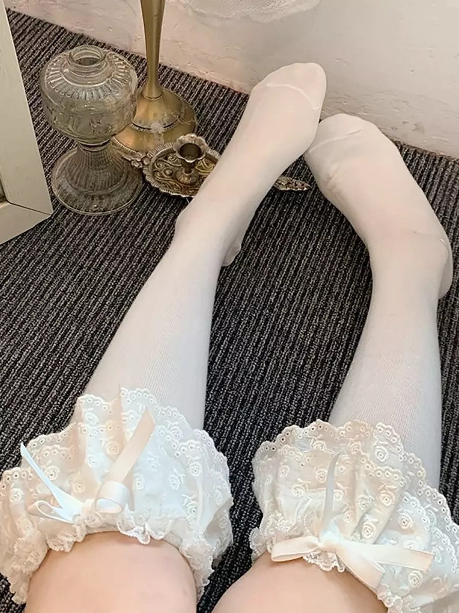 Close-up front-facing view of sweet lolita mid-calf socks showing white sock body with ruffled lace cuff and light-colored satin bow detail, barefoot on dark carpet
