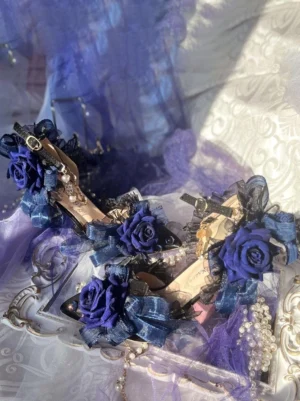 Pair of deep blue lolita stiletto sandals with ankle buckle straps and floral ribbon decorations, flat lay top-down view against damask fabric background with purple tulle and pearl strands
