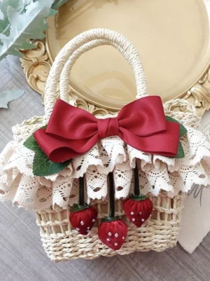 Lolita straw handbag with wine red satin bow and strawberry pendants, shot from elevated oblique angle on vintage wooden table with decorative tray and green foliage, showcasing lace ruffle trim and romantic pastoral aesthetic