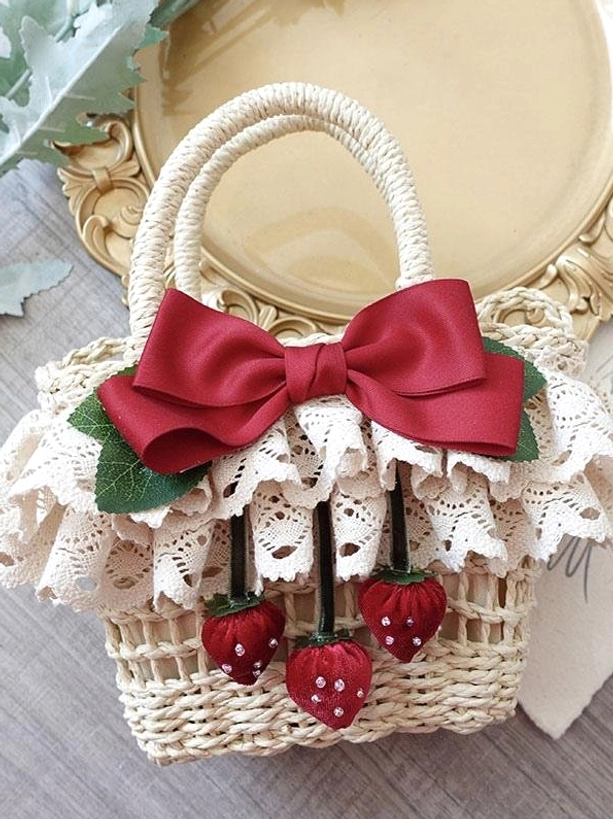 Lolita straw handbag with wine red satin bow and strawberry pendants, shot from elevated oblique angle on vintage wooden table with decorative tray and green foliage, showcasing lace ruffle trim and romantic pastoral aesthetic