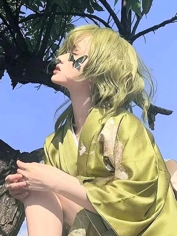 Green heat-resistant lolita wig with air bangs and wavy hair shown from high angle, sitting pose with decorative butterfly makeup