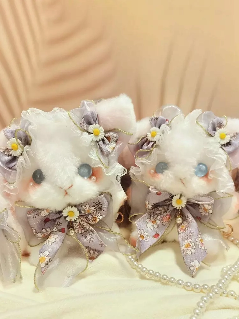Two matching white plush bunny crossbody bags displayed together on fabric tabletop with pearl chain, showing identical daisy bows and blue glass eyes