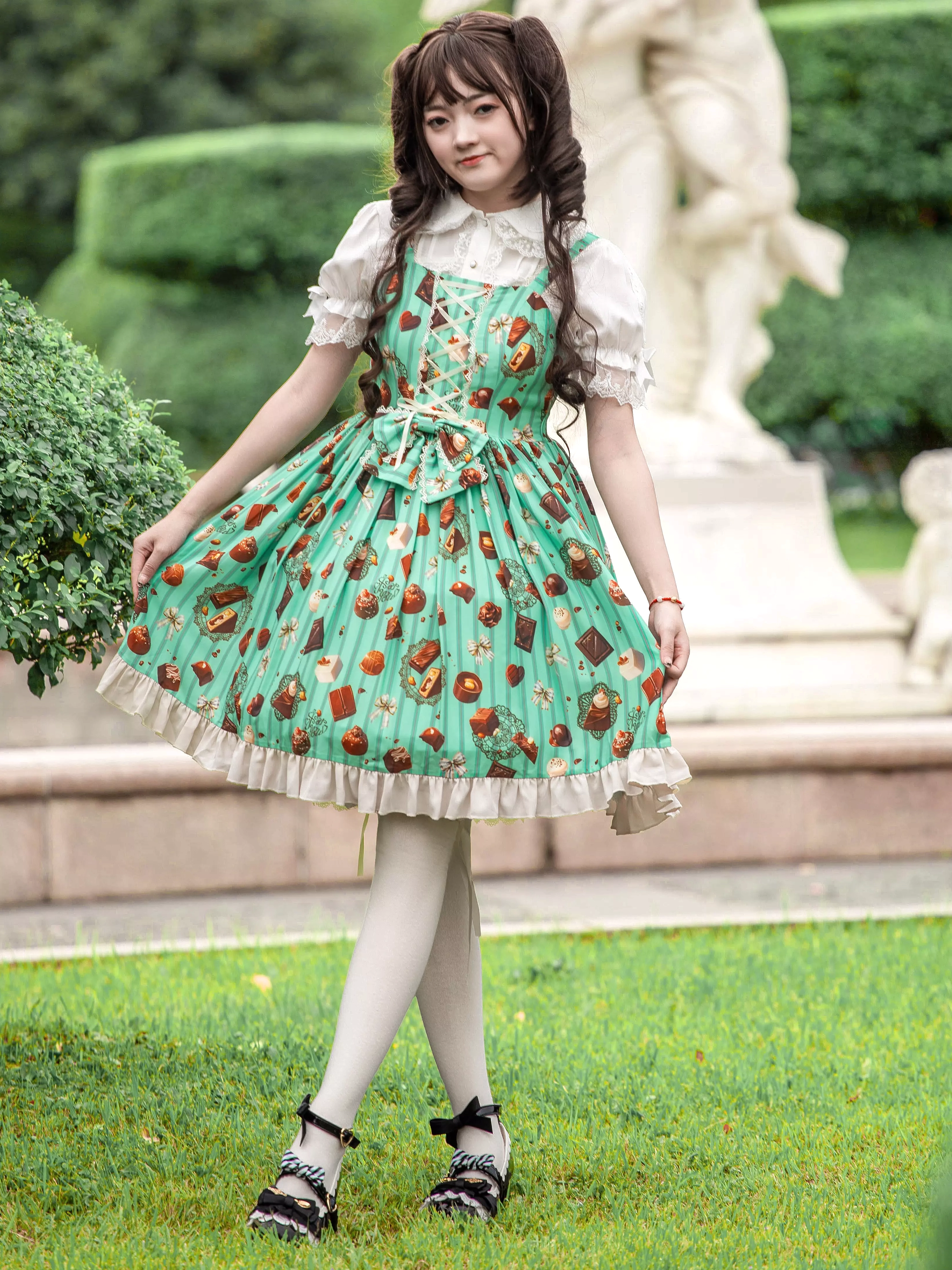 Side profile view of mint green chocolate print sweet Lolita JSK dress showing ruffled hem and white stockings garden stone statue background
