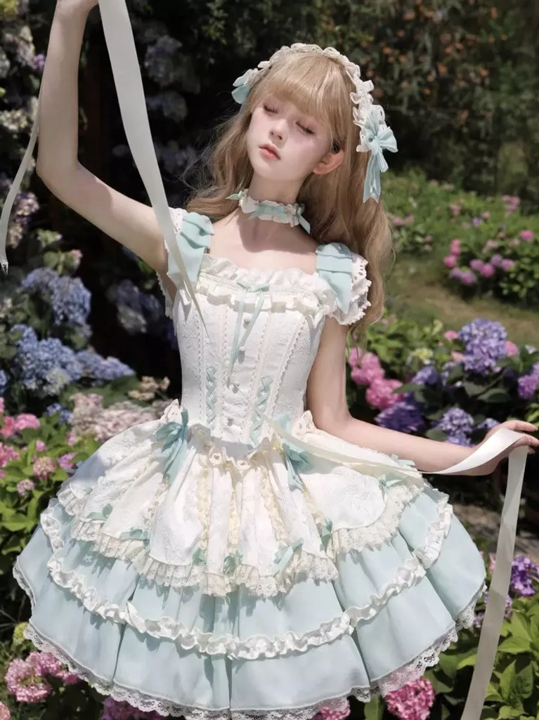 Mint green and off-white sweet lolita dress with lace-up front bodice, layered ruffles, and satin bows, worn by blonde model in outdoor flower garden setting, sleeveless design with front high-angle view